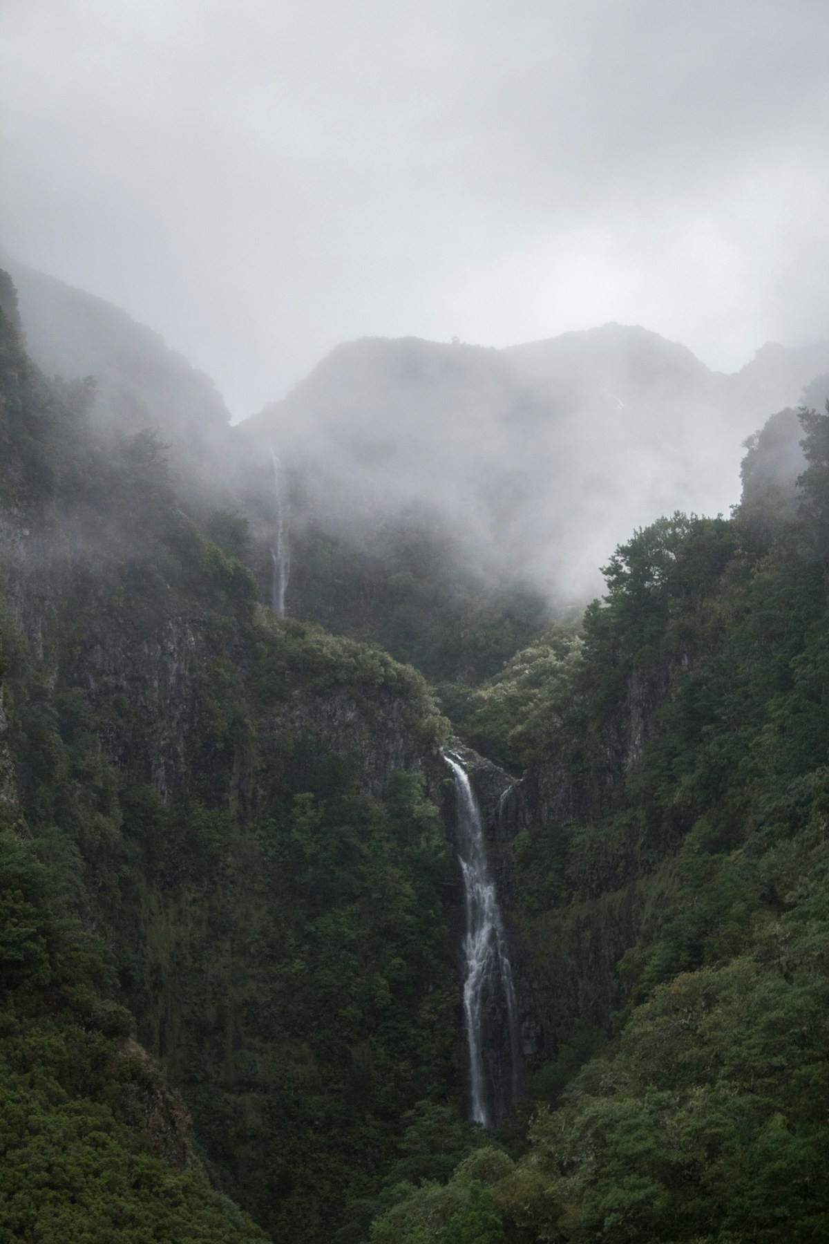 A tall waterfall cascades down mossy green volcanic cliffs in foggy weather at 25 Fontes Falls, Madeira, Portugal