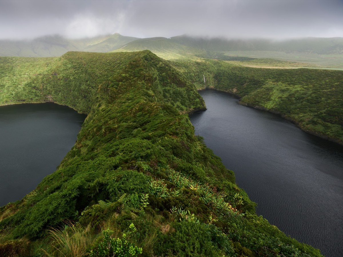 Twin dark crater lakes on Flores island, Azores, separated by a moss-covered ridge with a waterfall visible in the background