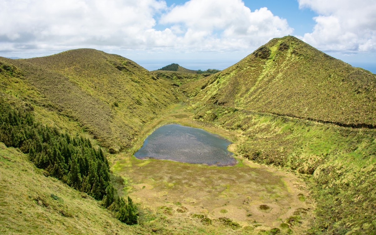 Small volcanic crater lake nestled between green volcanic hills on an Azores island, with the ocean visible in the distance