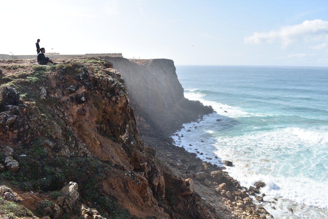 Two people sitting on the dramatic cliffs at Sagres with the Fortaleza visible in the background and waves breaking below
