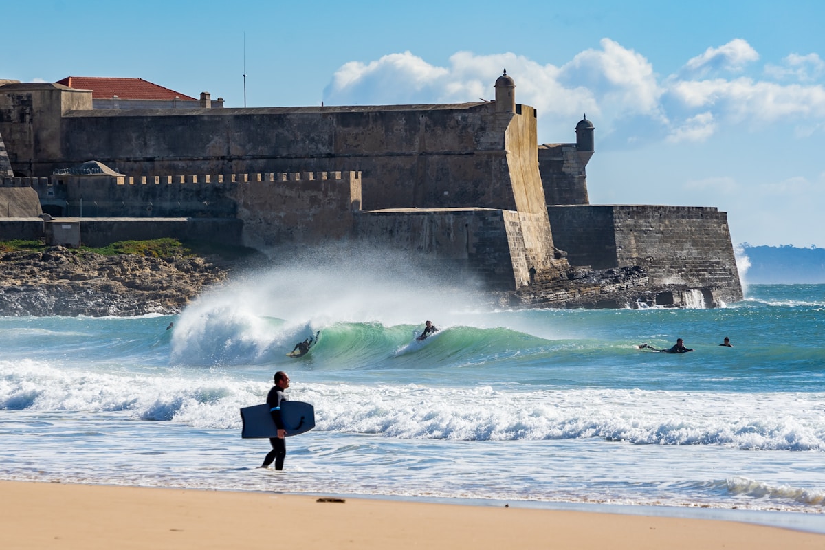 Surfers heading into Atlantic waves at Carcavelos beach with its 16th-century fortress behind, Lisbon coast