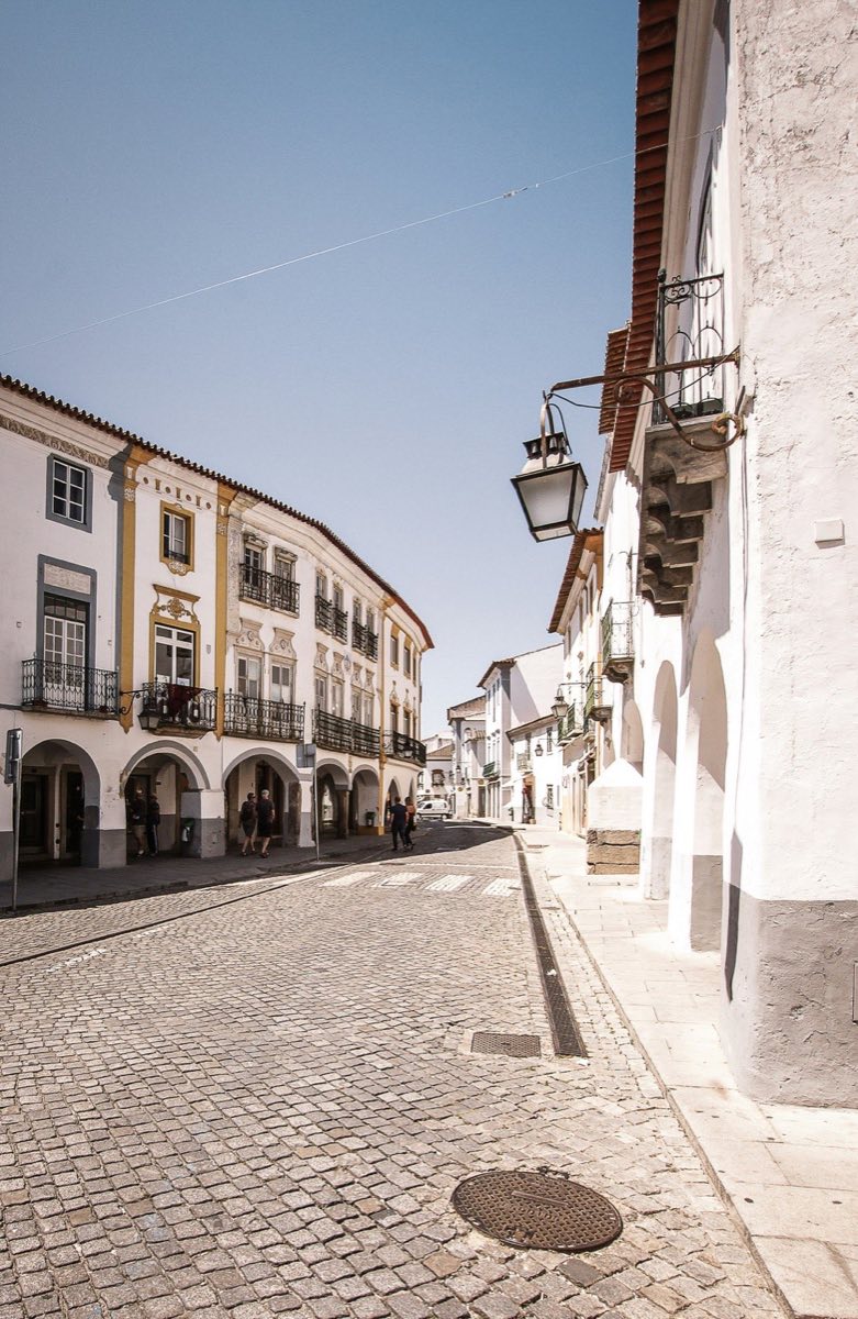 Cobblestone street in Évora lined with whitewashed Alentejo buildings under a clear blue sky, Portugal